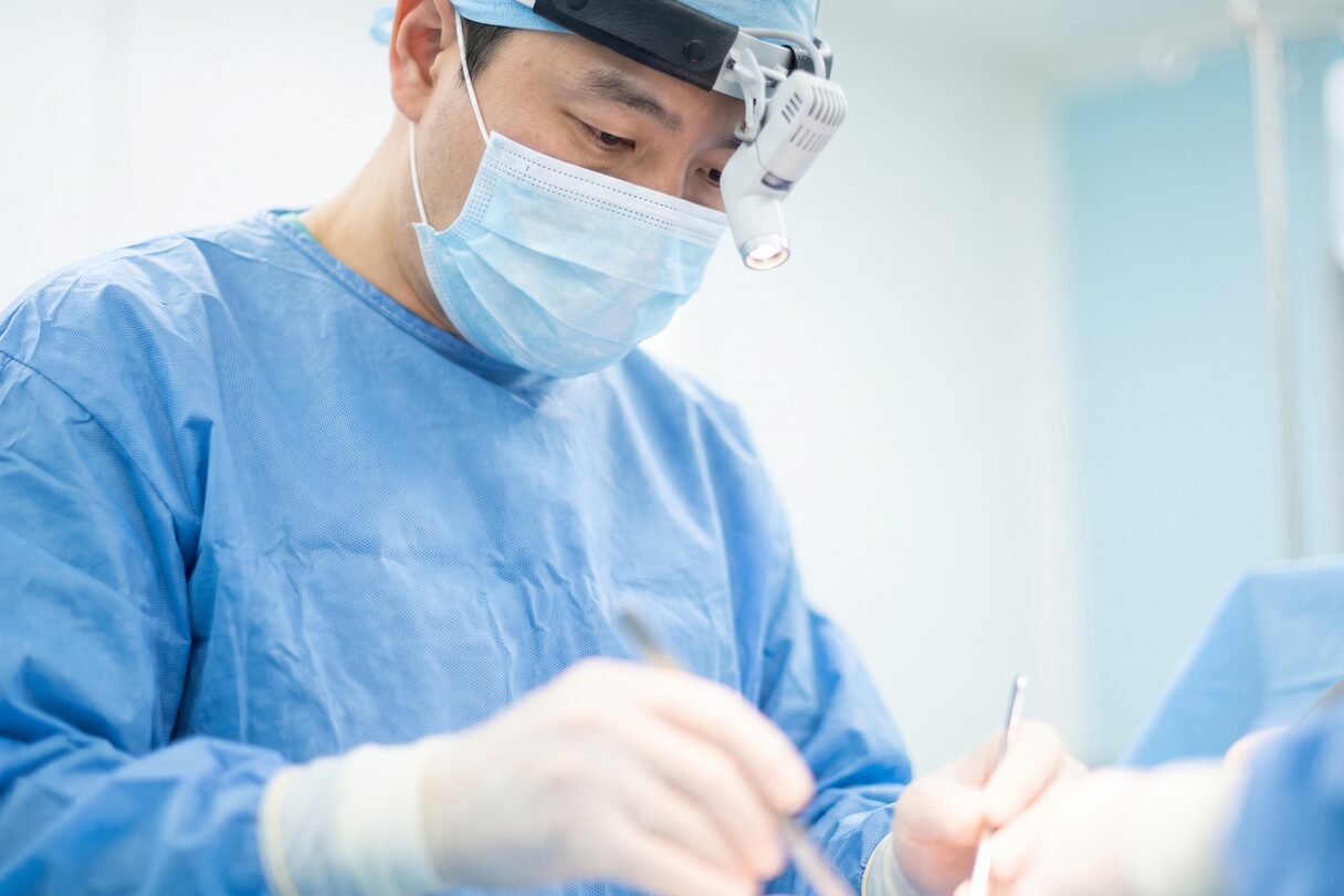 블루 수술 가운과 헤드램프, 마스크를 착용한 익명의 외과의가 시술에 집중하는 임상 컨셉 예시 이미지