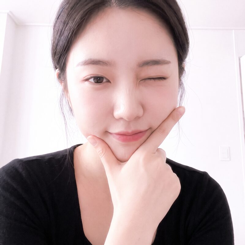 흰 벽 실내에서 한쪽 눈을 찡긋한 여성 방문객의 데일리 셀피 예시 이미지